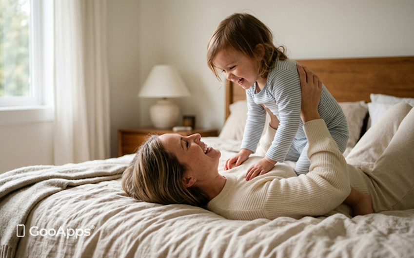 Fotografía íntima de una madre tumbada en una cama, levantando con alegría a su hijo pequeño hacia el aire. Ambos ríen y se miran con ternura en un dormitorio acogedor iluminado por una suave luz matinal. Los tonos son neutros y cálidos, transmitiendo una sensación de amor y conexión.